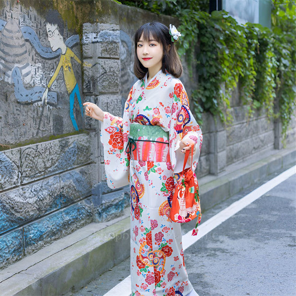 Jeune femme portant un kimono traditionnel japonais blanc avec des motifs floraux colorés. Elle porte un obi vert et un petit sac rouge. Kimonos à vendre.