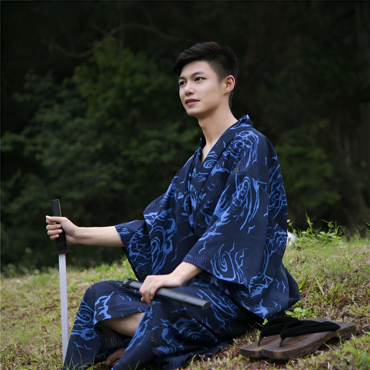 Homme asiatique portant un kimono bleu à motifs, assis dans la nature avec un katana et des sandales traditionnelles. Vêtements japonais authentiques en vente.
