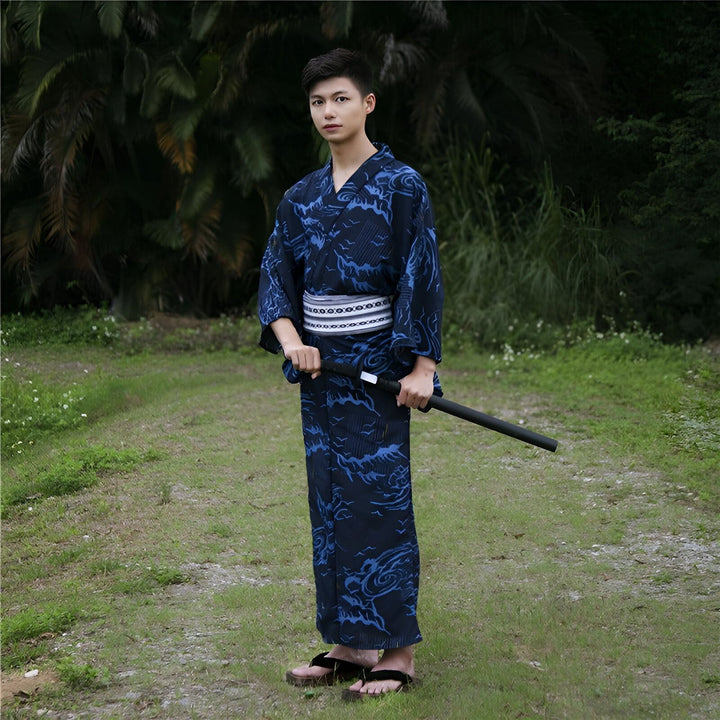 Jeune homme en kimono bleu marine avec motifs vagues, ceinture obi blanche et sandales traditionnelles. Il tient un sabre. Vêtements japonais traditionnels, yukata masculin. Boutique de kimonos authentiques.