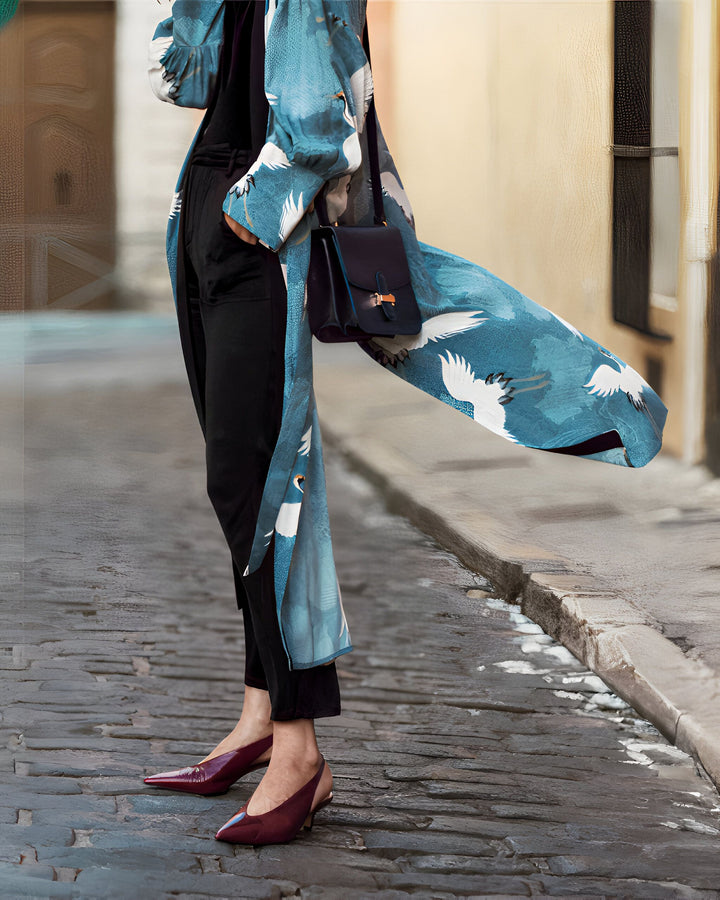 Femme portant un kimono bleu à motifs de grues blanches. Vêtements amples noirs, sac à main bandoulière et escarpins bordeaux complètent le look. Idéal pour un style élégant et décontracté. Découvrez notre collection de kimonos.