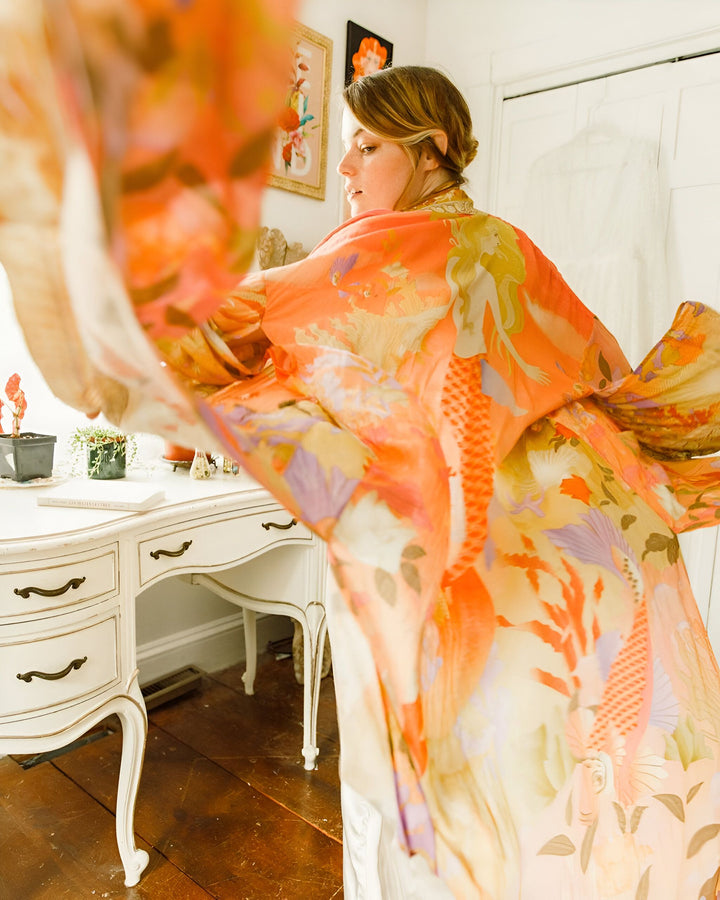 Femme portant un kimono orange à motifs devant un bureau vintage. Kimono ample et fluide, idéal pour se détendre avec style. Boutique de kimonos en ligne.