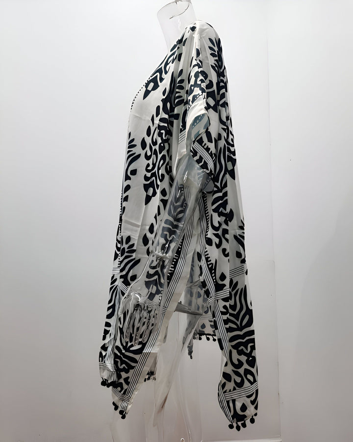 Kimono blanc à motifs noirs sur mannequin. Vêtement ample et léger, idéal pour l'été. Motifs originaux et finitions soignées avec des pompons. Découvrez notre collection de kimonos uniques!