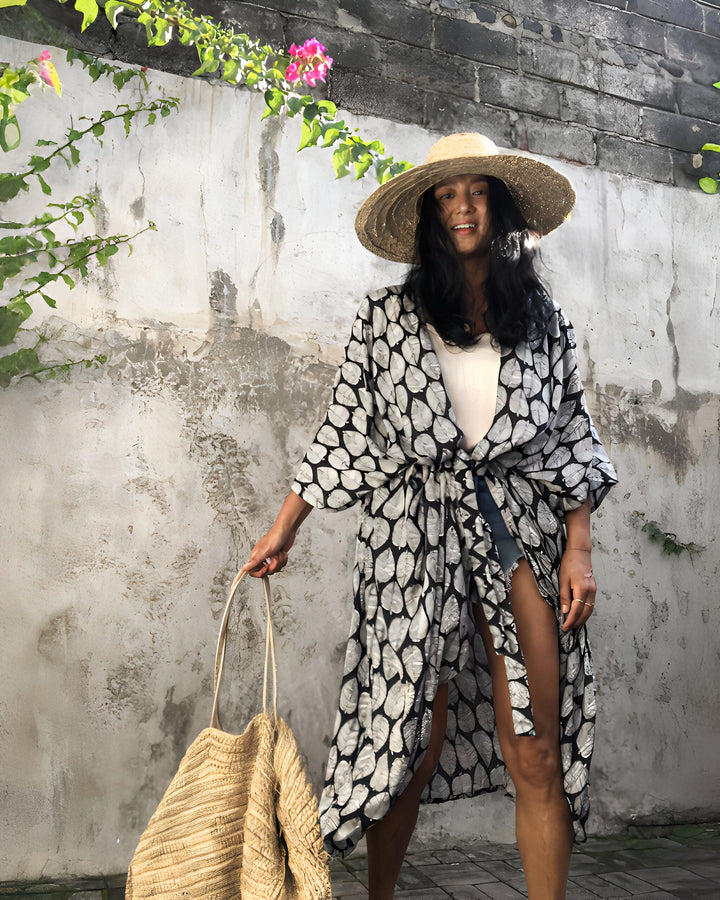 Femme souriante portant un kimono imprimé feuilles, chapeau de paille et sac en fibres naturelles. Mode décontractée et élégante. Découvrez notre collection de kimonos uniques.
