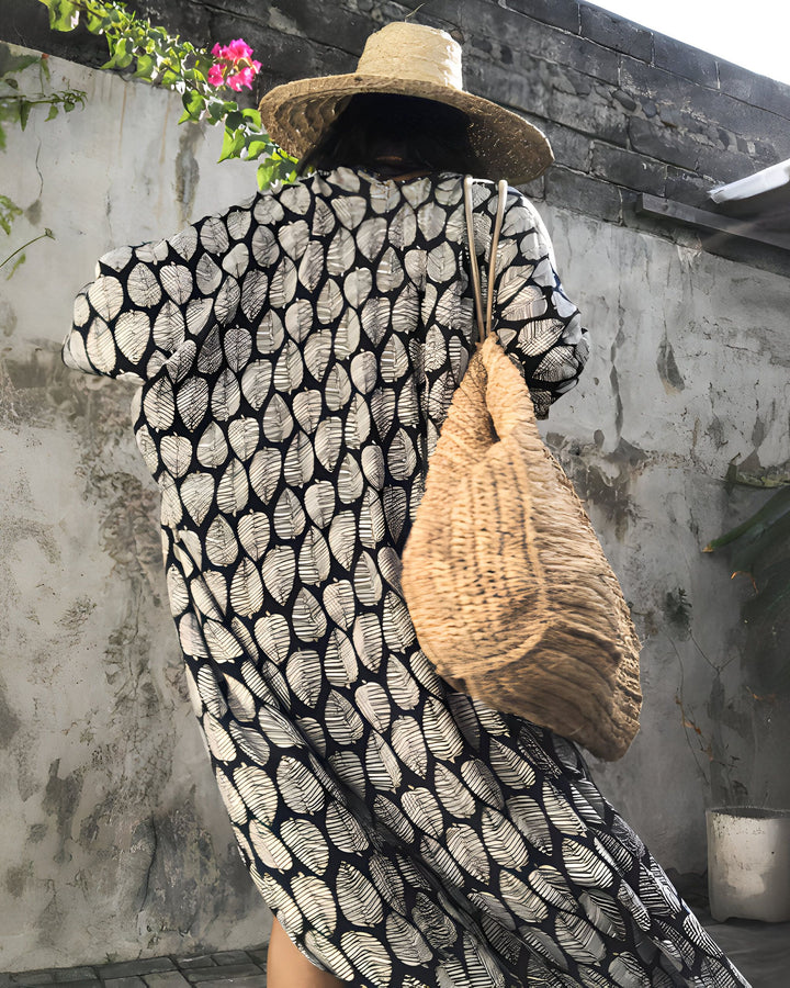Femme portant un kimono à motifs de feuilles noir et blanc, avec un chapeau de paille et un sac en fibres naturelles. Kimonos élégants et confortables en vente.