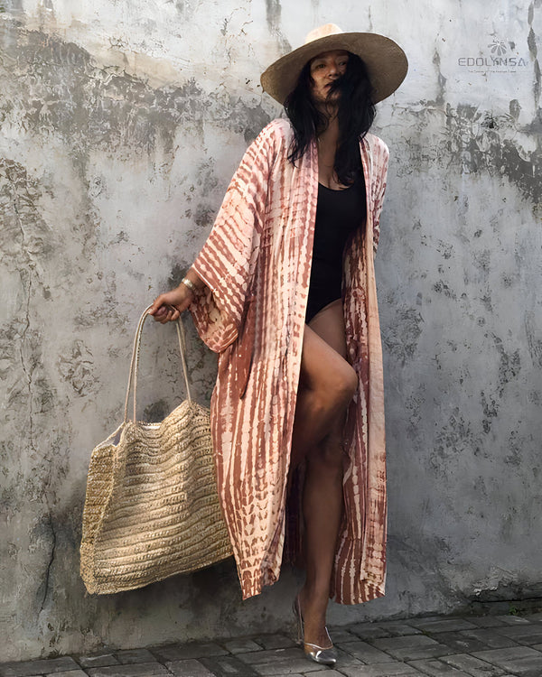 Femme en kimono tie-dye rose et blanc, maillot de bain noir, chapeau de paille et talons argentés. Elle porte un sac de plage en paille devant un mur en béton texturé.