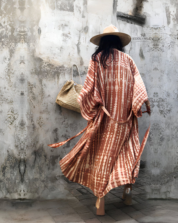 Femme vue de dos portant un kimono tie-dye marron, un chapeau de paille et un sac en raphia. Elle marche devant un mur texturé en béton gris.