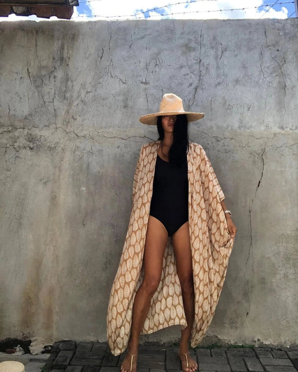 Femme portant un kimono beige à motifs de feuilles, un maillot de bain noir et un chapeau de paille. Idéal pour un look de plage élégant. Découvrez notre collection de kimonos.