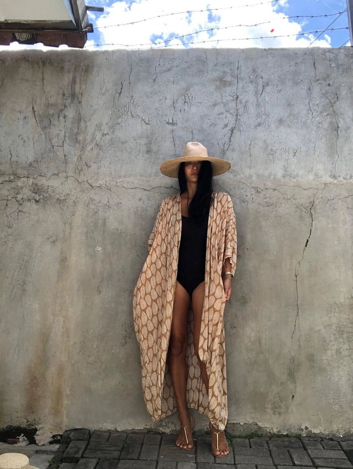Femme portant un kimono beige à motifs feuilles, maillot de bain noir et chapeau de paille. Vente de kimonos élégants et confortables pour toutes les occasions.