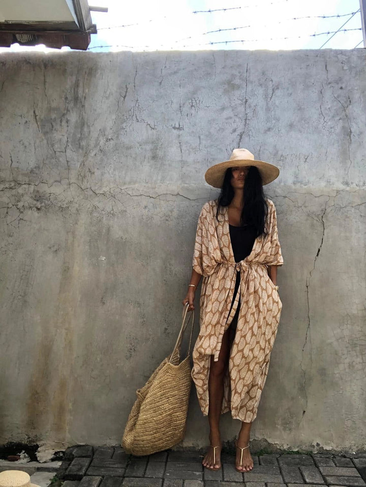 Femme portant un kimono marron à motifs, chapeau de paille et sac en osier. Look décontracté pour la plage ou la ville. Découvrez notre collection de kimonos!