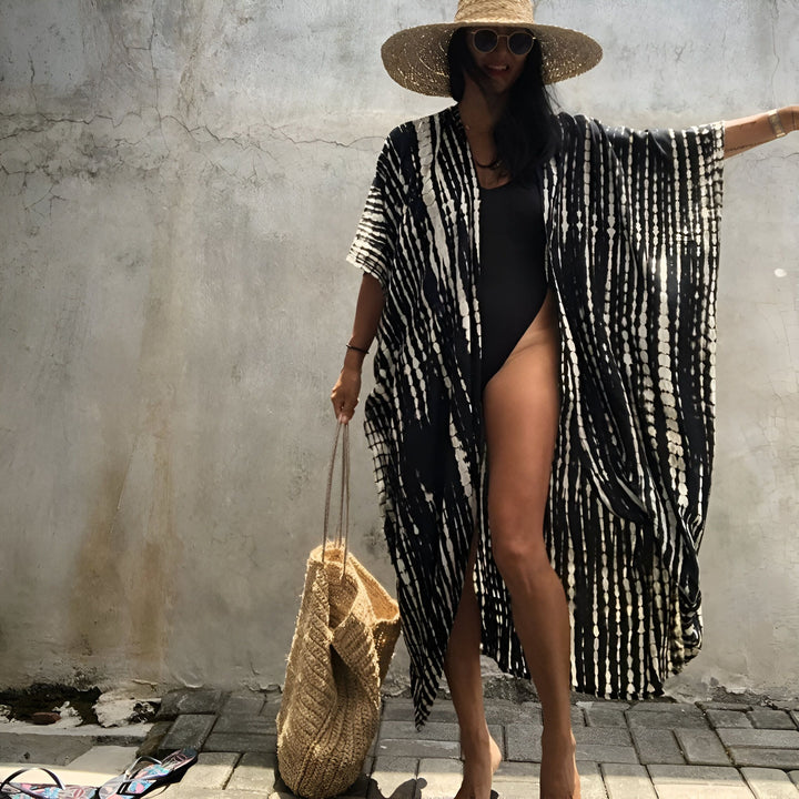 Femme portant un kimono noir et blanc sur un maillot de bain noir, avec un chapeau de paille et un sac de plage en paille. Vente de kimonos en ligne.