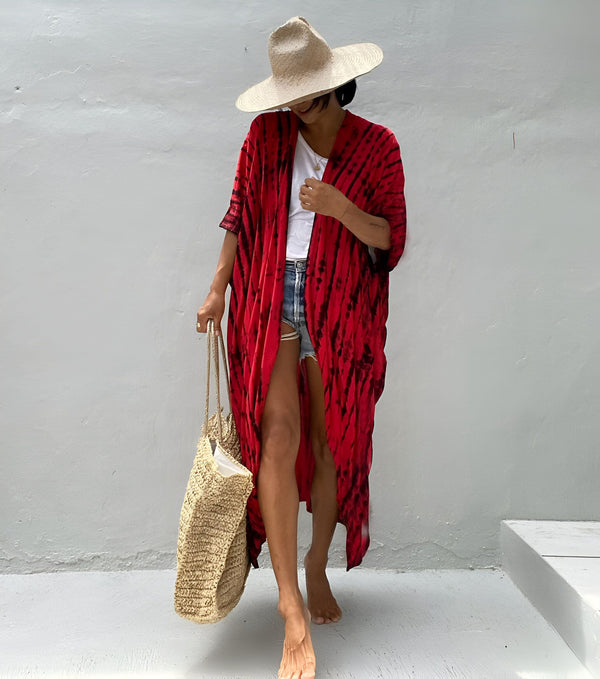 Femme portant un kimono rouge tie-dye, un short en jean, un chapeau de paille et un sac de plage en osier. Kimonos tendance pour femmes.