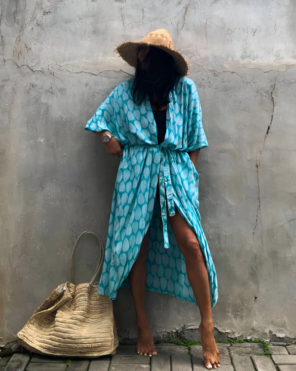 Femme portant un kimono turquoise à motifs, un chapeau de paille et un sac en raphia. Fond de mur en béton texturé. Look estival décontracté.