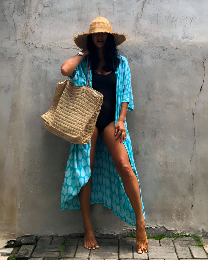 Femme en maillot de bain noir avec paréo turquoise et chapeau de paille, tenant un grand sac de plage en fibres naturelles. Pose décontractée devant un mur texturé. Look estival et décontracté pour les vacances à la plage. Style bohème et chic. Mode plage.
