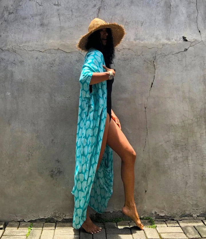 Femme souriante portant un chapeau de paille et un kimono turquoise sur un maillot de bain noir devant un mur en béton texturé. Elle se tient pieds nus sur un chemin pavé. Style de plage décontracté.
