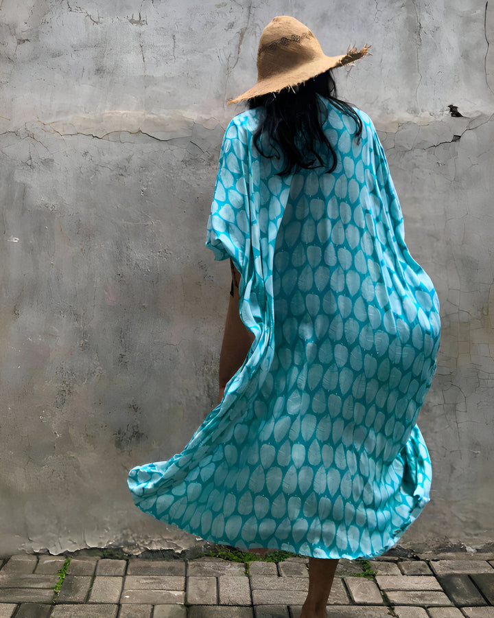 Une femme portant un chapeau de paille et un kimono turquoise à motifs de feuilles, dos à la caméra, devant un mur texturé.