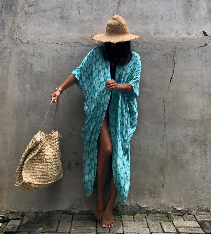 Femme en kimono turquoise, chapeau de paille et sac en osier devant un mur gris. Style bohème, vacances d'été, mode plage, accessoires naturels.