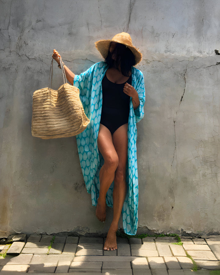 Femme en maillot de bain noir, kimono bleu à motifs, chapeau de paille et sac de plage. Elle se tient adossée à un mur gris texturé. Ambiance estivale et décontractée. Vacances à la plage. Style bohème. Accessoires de mode.
