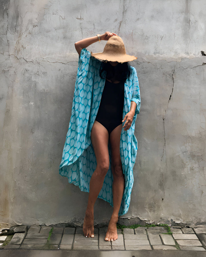 Femme en maillot de bain noir et kimono turquoise à motifs, chapeau de paille, posant devant un mur texturé. Mode estivale, style de plage décontracté, look vacances ensoleillées.