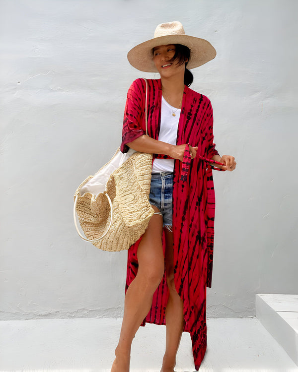 Femme souriante avec chapeau de paille et kimono rouge tie-dye. Elle porte un short en jean, un t-shirt blanc et un sac en paille. Idée de look décontracté et estival.