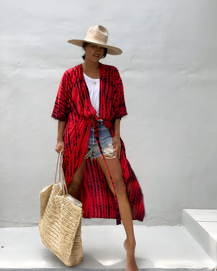 Femme souriante portant un chapeau de paille, un kimono rouge et noir, un short en jean et un sac de paille. Style estival décontracté.