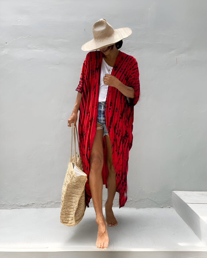 Femme pieds nus portant un kimono rouge et noir tie-dye, un short en jean, un t-shirt blanc et un chapeau de paille. Elle porte un sac de plage en paille et se tient devant un mur blanc texturé. Style décontracté et estival.