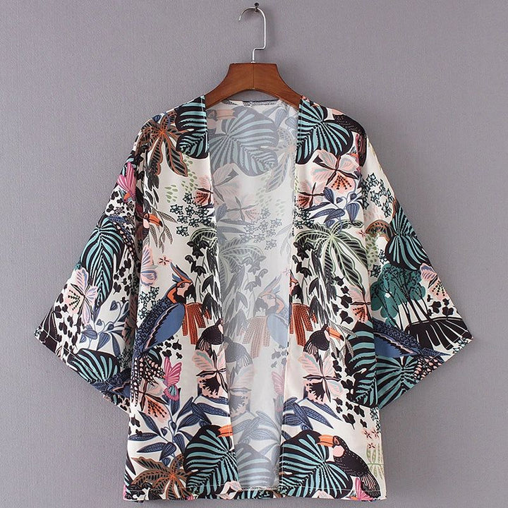 Kimono pour femme à motifs floraux et tropicaux suspendu à un cintre en bois. Vêtement d'été léger et coloré avec des feuilles de palmier et des toucans. Mode féminine, style bohème.