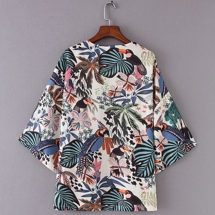 Kimono à motifs de toucans et de feuilles tropicales, suspendu à un cintre en bois. Vue de dos, manches larges. Tissu blanc avec imprimé coloré. Mode estivale, style décontracté.