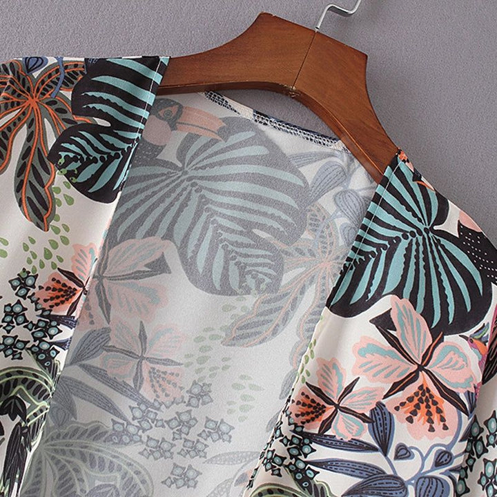 Gros plan sur un kimono à motifs floraux tropicaux, suspendu à un cintre en bois. Détail du col en V et des imprimés de feuilles et fleurs dans les tons de bleu, rose et orange. Idéal pour un look estival et décontracté.