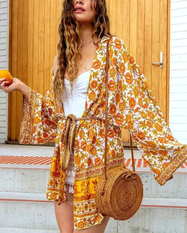 Femme portant un kimono fleuri jaune et orange, tenant une orange. Kimono d'été avec imprimé floral, idéal pour un look bohème chic. Vêtements style kimono à la mode.