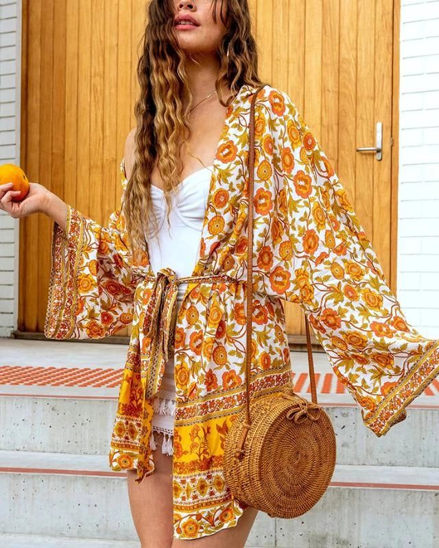 Femme portant un kimono fleuri jaune et orange, tenant une orange. Kimono d'été avec imprimé floral, idéal pour un look bohème chic. Vêtements style kimono à la mode.
