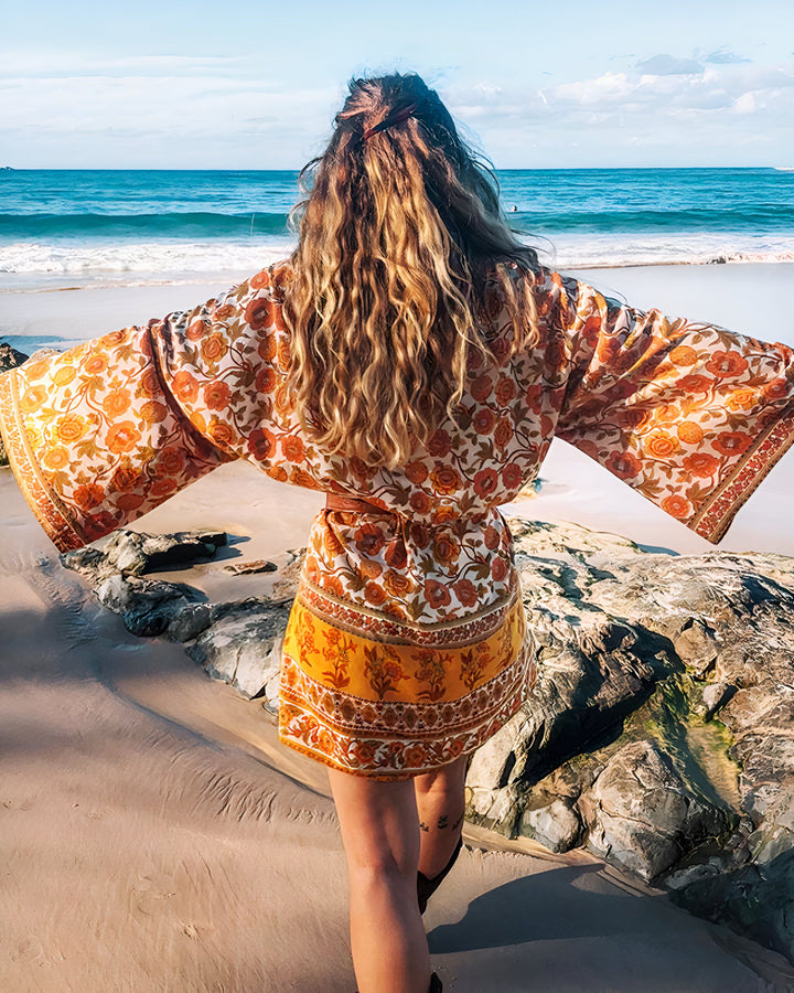 Femme vue de dos en kimono fleuri, bras ouverts face à la mer. Kimonos bohèmes pour un style unique. Collection de kimonos imprimés pour toutes les occasions. Mode femme, vêtements de plage, idée cadeau. Achetez maintenant !