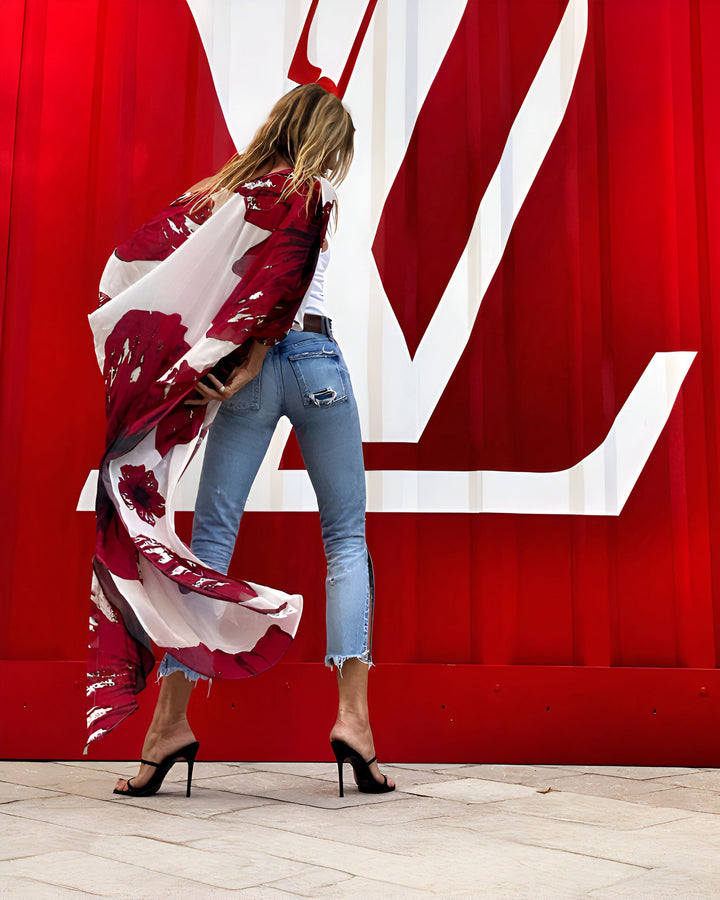 Une femme vue de dos, en jean et kimono fleuri rouge et blanc, pose devant un fond rouge orné d'un grand logo blanc. Elle porte des talons hauts noirs et semble marcher avec élégance. Mode et style.