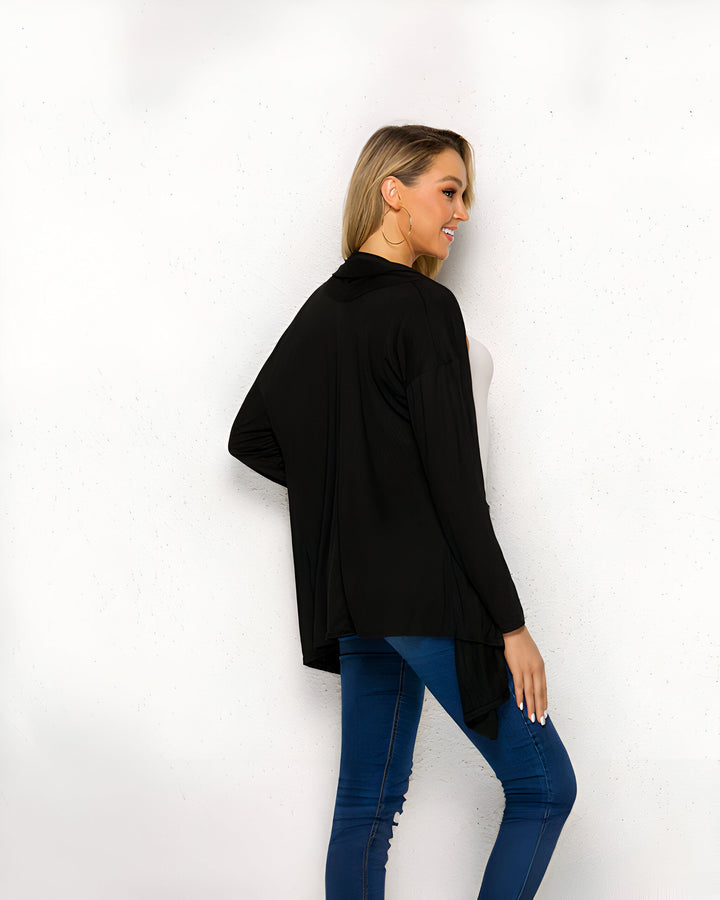 Femme blonde souriante, vue de dos, portant un cardigan noir fluide sur un débardeur blanc et un jean bleu moulant. Elle porte des boucles d'oreilles dorées.