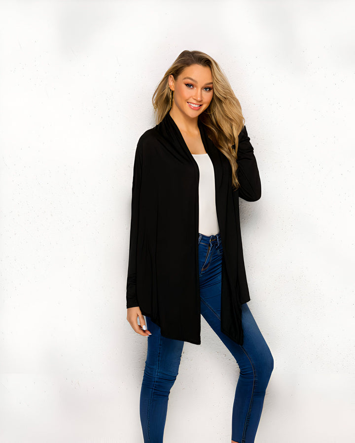 Une femme blonde souriante porte un cardigan noir ouvert sur un débardeur blanc et un jean bleu devant un fond blanc texturé.