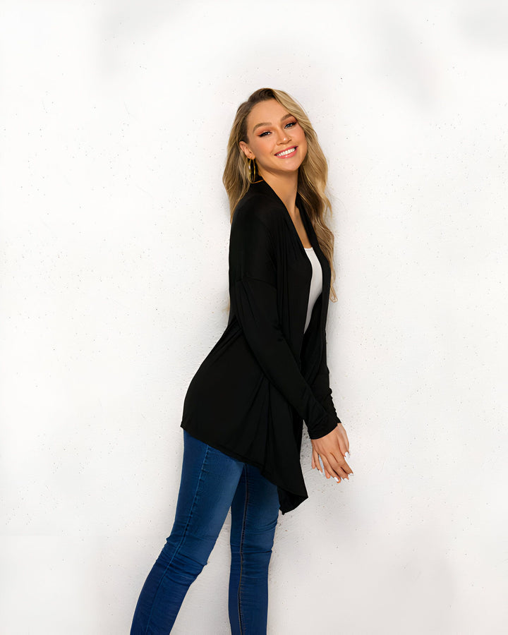 Une femme souriante porte un cardigan noir ample sur un débardeur blanc, avec un jean bleu. Elle pose devant un mur blanc texturé. Ses longs cheveux blonds ondulés et ses boucles d'oreilles dorées complètent son look décontracté et élégant.