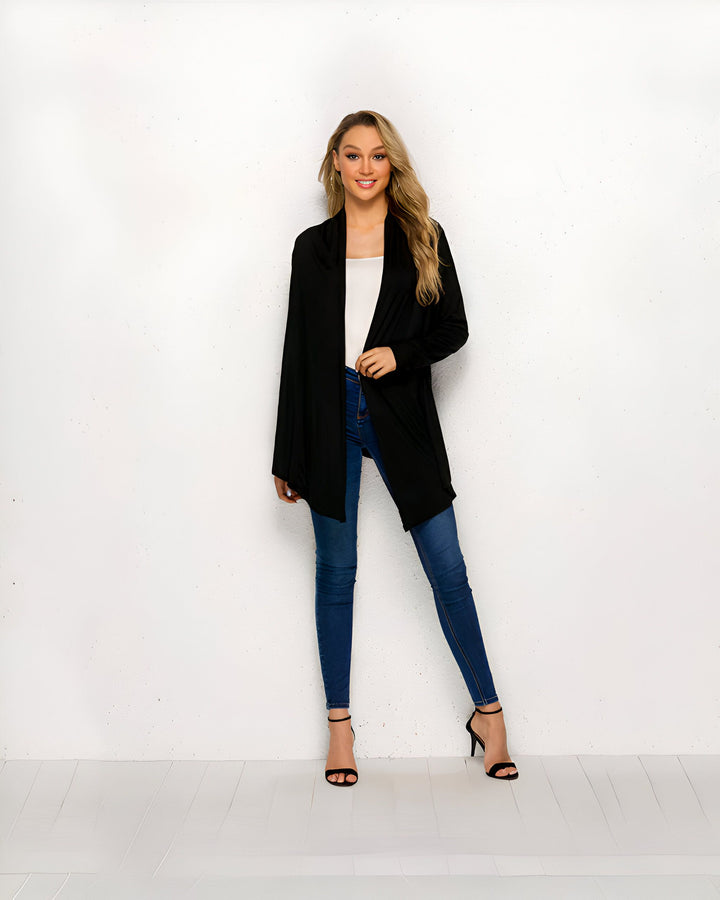 Une femme blonde souriante portant un cardigan noir long, un débardeur blanc, un jean skinny et des talons noirs sur un fond blanc. Vêtement décontracté et élégant.
