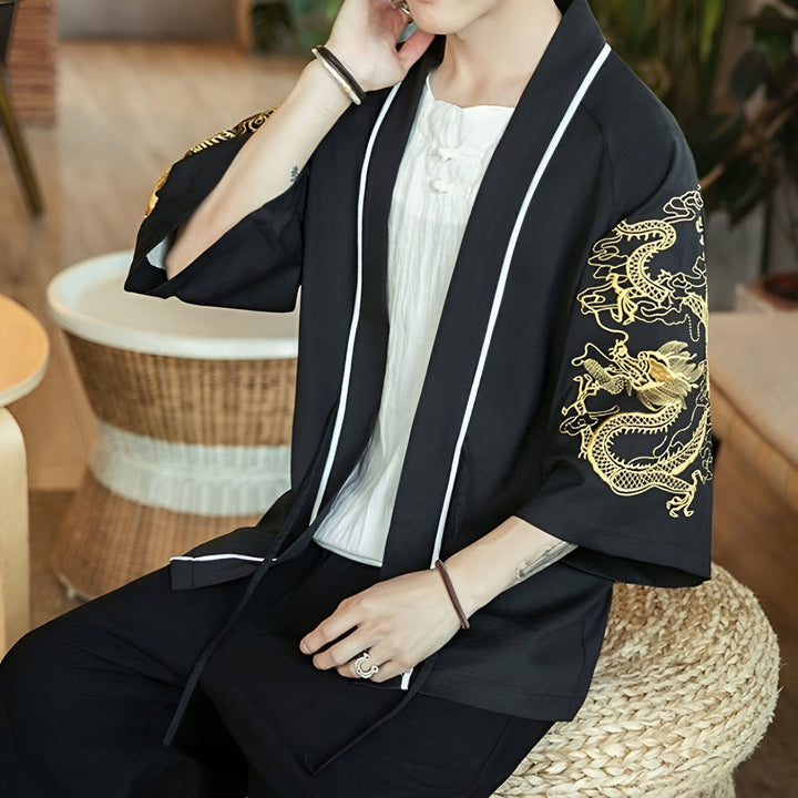 Un homme portant un kimono noir avec des broderies de dragons dorés sur les manches. Il porte une chemise blanche à col mao en dessous et est assis sur un tabouret en osier.