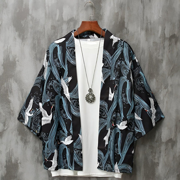 Kimono noir imprimé avec des grues blanches et motifs vagues bleues, porté sur un t-shirt blanc avec un collier, suspendu à un cintre devant un fond gris texturé. Vêtement d'extérieur à la mode.