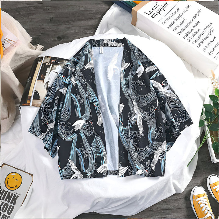 Kimono noir à motifs de grues blanches et vagues bleues, posé sur un drap blanc. Chaussures jaunes et sac en papier en arrière-plan. Style japonais décontracté.