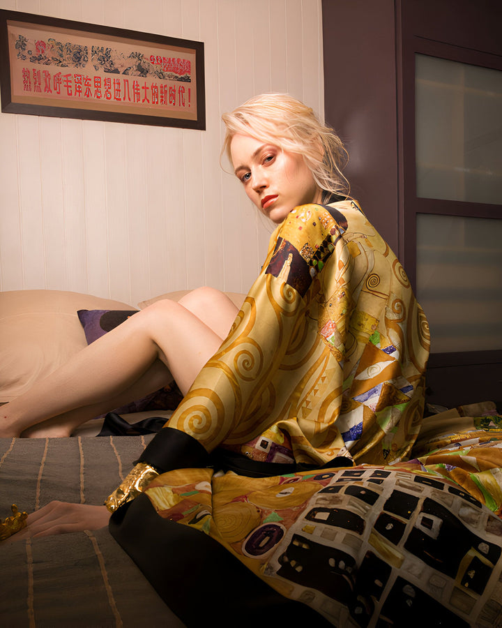 Une femme blonde aux yeux bleus, assise sur un lit, porte un kimono doré inspiré de Klimt. Une affiche chinoise est accrochée au mur au-dessus du lit. Ambiance intime et artistique.