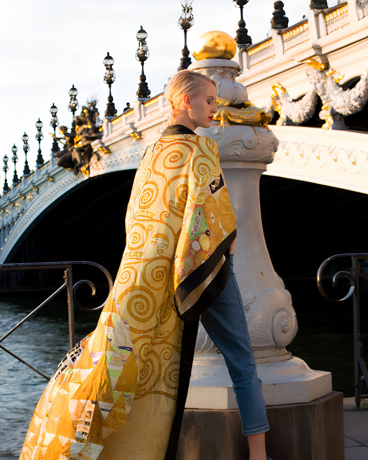 Une femme blonde, vue de dos, porte un kimono doré à motifs tourbillonnants sur le pont Alexandre-III à Paris. L'architecture ornementale du pont, avec ses dorures et ses lampadaires, est visible en arrière-plan.