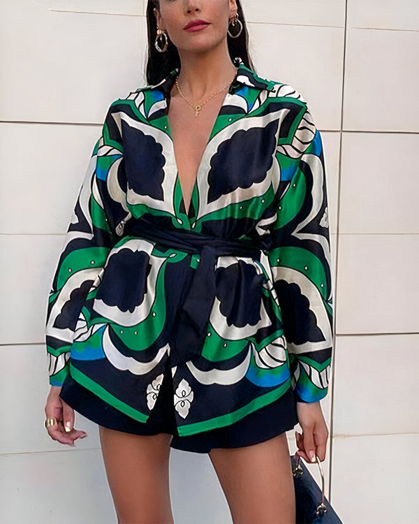 Femme portant un ensemble short et veste à motifs verts, bleus et noirs avec une ceinture noire. Elle porte des boucles d'oreilles et un collier dorés.