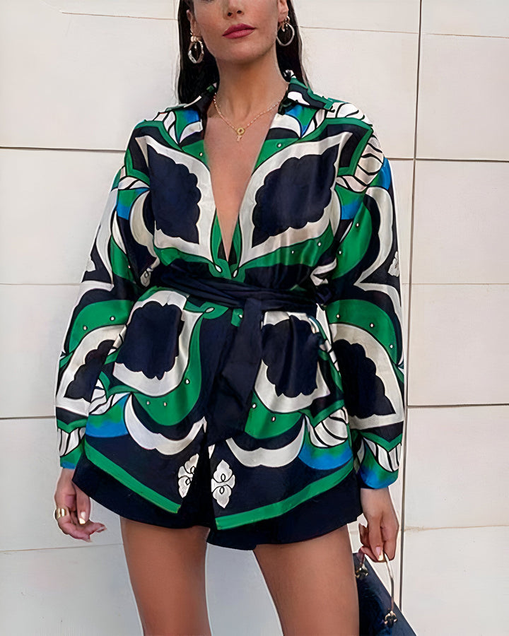 Femme portant un ensemble short et veste à motifs verts, bleus et noirs avec une ceinture noire. Elle porte des boucles d'oreilles et un collier dorés.