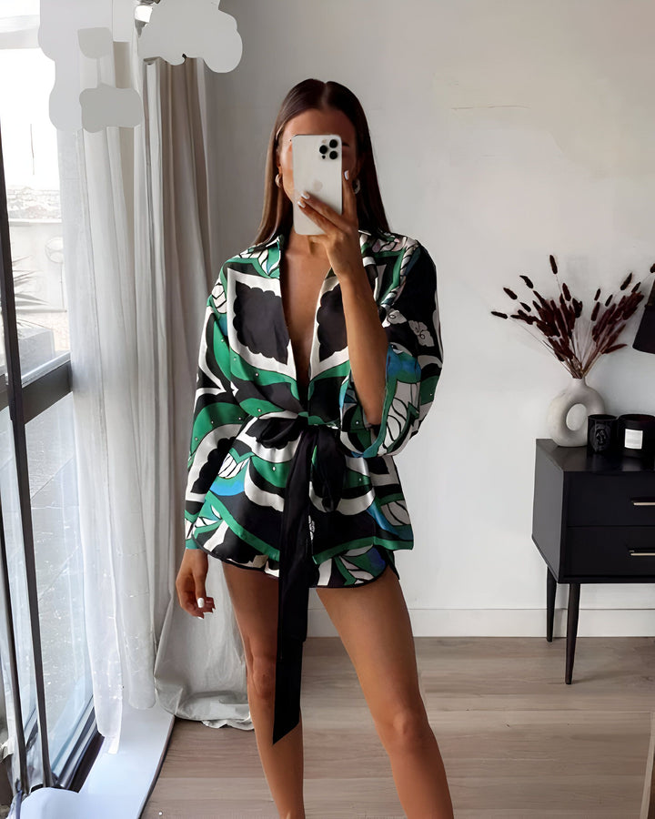 Femme prenant un selfie en pyjama chic à motifs verts, noirs et blancs, noué à la taille avec une ceinture noire. Arrière-plan intérieur moderne avec fenêtre et décoration.