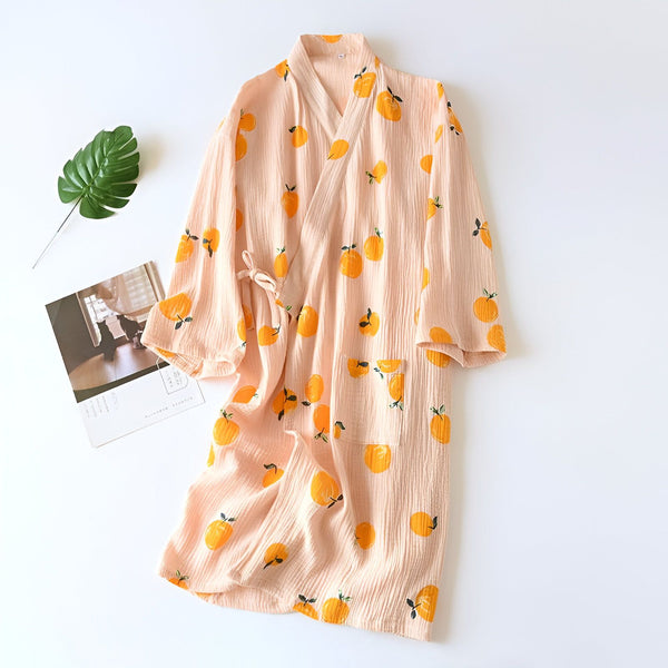 Peignoir rose à motifs d'agrumes, style kimono. Vue rapprochée du peignoir avec ceinture nouée, poche plaquée, feuille verte et magazine. Vêtement de nuit confortable et élégant.