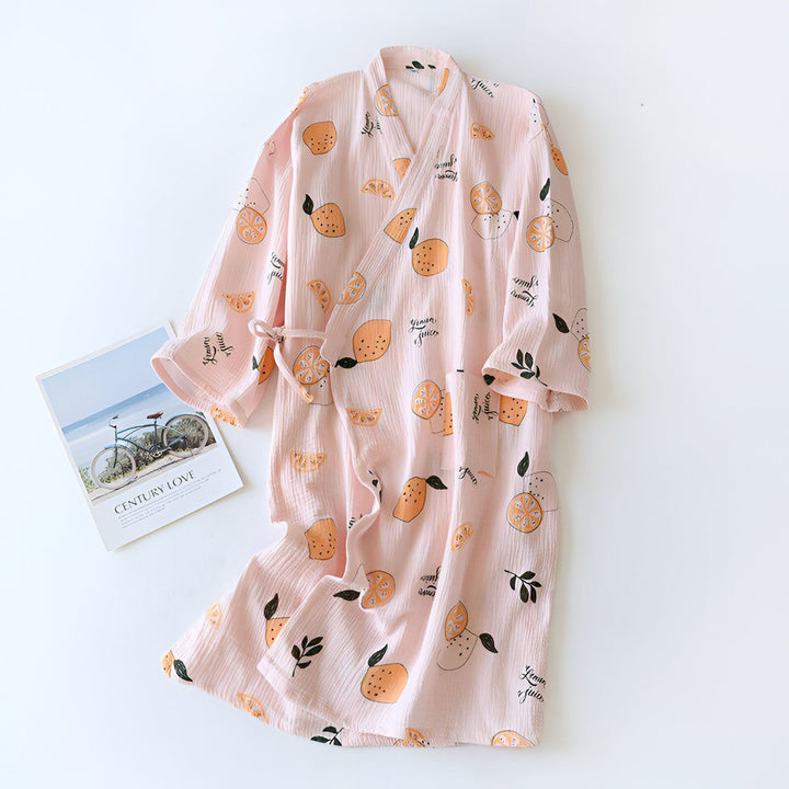 Peignoir rose à motifs d'agrumes et feuilles noires, style kimono avec ceinture, posé sur une surface blanche, avec un livre intitulé "Century Love" à côté. Vêtement confortable, léger et estival.