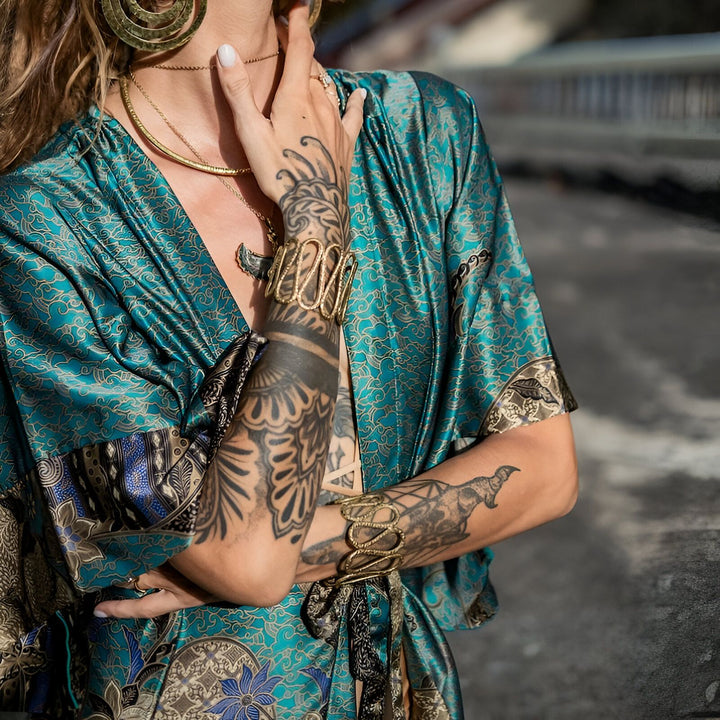 Femme portant un kimono en soie turquoise à motifs, ornée de bijoux dorés (colliers, boucles d'oreilles, bracelets). Tatouages visibles sur les bras et les mains. Vêtements kimono élégants et confortables.