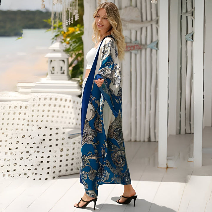 Une femme blonde porte un kimono bleu et blanc à motifs cachemire sur un haut court blanc et un pantalon blanc. Elle se tient sur une terrasse en bois avec vue sur l'océan. Ses cheveux blonds bouclés tombent sur ses épaules.