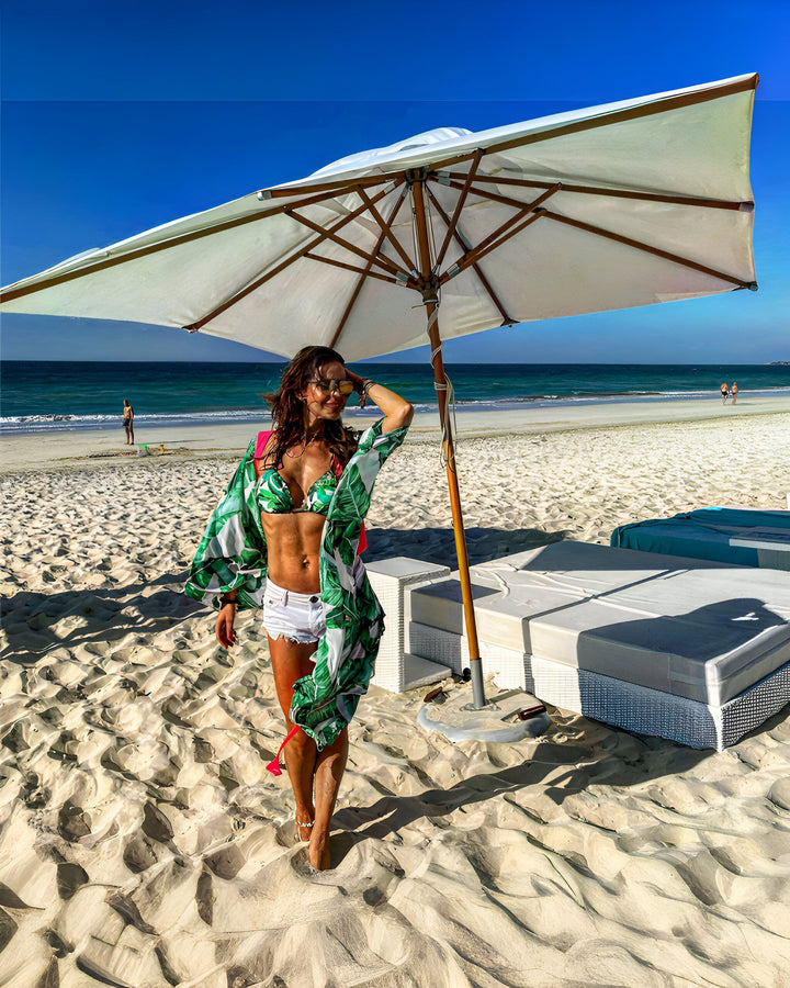 Femme en bikini et paréo sur plage de sable blanc, sous parasol. Mer turquoise et ciel bleu en arrière-plan. Chaises longues confortables. Ambiance vacances et détente au bord de l'eau.