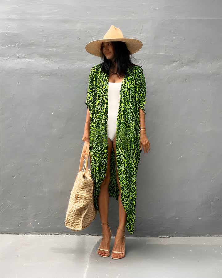 Femme portant un kimono vert à imprimé léopard, un maillot de bain blanc et un chapeau de paille. Sac de plage tressé. Style estival, mode kimono.